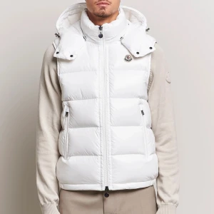 Moncler väst - Säljer en snygg vit dunväst från Moncler i storlek 2XL. Västen har hög krage, avtagbar huva och två dragkedjefickor framtill. Klassisk Moncler-logga på bröstet och quiltad design. Perfekt för lager-på-lager under kyliga dagar.