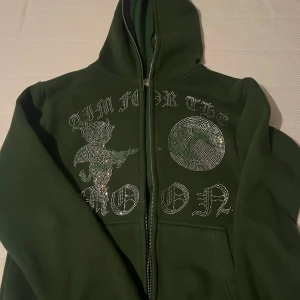 Grön hoodie med strasstenar och tryck - Mörkgrön hoodie med huva och dragkedja. Framsidan har ett stort tryck med texten 'AIM FOR THE MOON' och motiv av en figur och måne i glittriga strasstenar. Praktiska fickor framtill och långärmad modell. Perfekt för dig som gillar streetwear med extra bling.