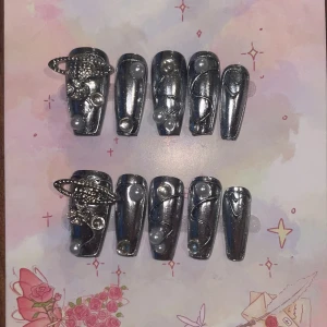 M - Kromade lösnaglar med pärlor och planetdekor - Snygga lösnaglar i spegelblank krom med silverfinish. Dekorerade med vita pärlor och tunna svarta linjer, samt coola 3D-planetdetaljer i metall på två naglar. Naglarna är långa och har en rak, kistformad design. Perfekt för dig som vill sticka ut med futuristisk stil.