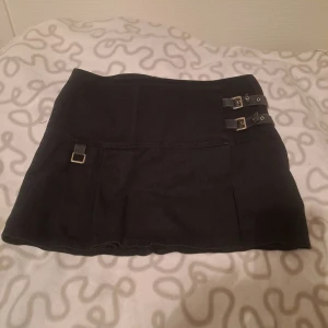 Svart kortkjol med spännen - Svart kortkjol i jeansmaterial med två coola spännen och metallöglor på sidan. Plisseringar vid sidan framtill. 🩷😊 Str 38