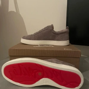 Grå mocka sneakers från Louboutin oanvända helt nya. - Snygga sneakers från Christian Louboutin i grå mocka med klassisk vit sula och den ikoniska röda undersidan. Skorna har rund tå, snörning och diskreta metalldetaljer vid sidan. Perfekta för dig som vill ha en stilren och lyxig look.