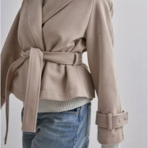 Beige kort omlott kappa med bälte - Säljer denna populära korta kappan från Nelly  med omlott-design och bälte i midjan. Jackan har breda slag, långa ärmar med snygga manschetter och knappar. Materialet är mjukt och känns som ull eller syntet. Perfekt för dig som gillar stilrena och trendiga plagg. Köpt för nypris 799 men säljer för 500 pris kan diskuteras.💓