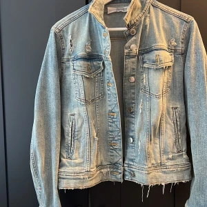Ljusblå jeansjacka med slitningar - Säljer en ljusblå jeansjacka från Denim med coola slitningar och rå kant nertill. Jackan har klassiska bröstfickor med lock, knappar framtill och långa ärmar. Perfekt för dig som gillar en avslappnad och trendig stil.