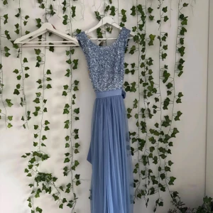 Ljusblå långklänning från Maya Petite - Säljer en ljusblå långklänning från Maya Petite med glittrig paljett-topp och omlott-detalj i ryggen. Kjolen är i mjukt, transparent tyg med volang och ett brett knytband i midjan. Perfekt för dig som vill sticka ut med en elegant look.