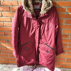 Cellbes vinröda jacka, storlek 38/40 - inröd jacka med dragkedja och knappar. Jackan har en avtagbar huva med fuskpäls. Det finns två fickor fram med dragkedja och tryckknapp. Jackan har även en innerficka.