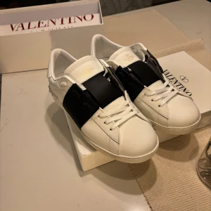 Valentino Garavani Open - Säljer ett par snygga Valentino Garavani Open sneakers i vitt skinn med bred svart läderdetalj över mitten. Klassisk rund tå, vita snören och diskret guldlogga på plösen. Kommer med originalkartong och dustbag. Perfekta för dig som gillar stilrena och lyxiga sneakers. Pris ej hugget i sten