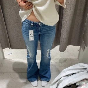 Lågmidjade bootcut jeans med slitningar - Säljer ett par blå lågmidjade bootcut jeans med snygga slitningar på framsidan. Jeansen har låg midja, klassiska fem fickor och coola detaljer på bakfickorna. Perfekta för dig som gillar en avslappnad och trendig stil.