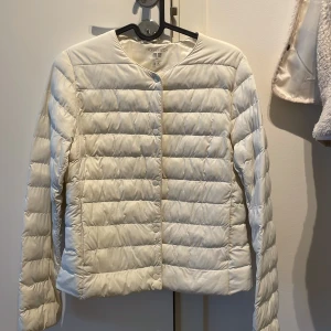 Vit pufferjacka från Uniqlo XXS - Lätt och stilren vit pufferjacka från Uniqlo i storlek XXS. Jackan har rund hals, tryckknappar framtill och quiltad design. Tillverkad i polyamid och polyester, perfekt för lager-på-lager under kyliga dagar.