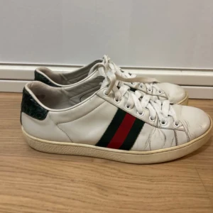 Gucci ace - Snygga vita sneakers från Gucci med klassiska röd och grön rand på sidan och mörkgrön detalj vid hälen. Skorna har snörning och är tillverkade i skinn. Perfekta för dig som vill ha en stilren och lyxig look med en ikonisk touch.