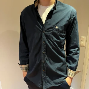 Mörkblå Burberry skjorta i bomull - Snygga mörkblå skjorta från Burberry med klassiskt rutigt tyg på insidan av kragen och manschetterna. Skjortan har knappar framtill, bröstficka med Burberry-logga och är tillverkad i 100% bomull. Perfekt för dig som gillar stilrena och tidlösa plagg.