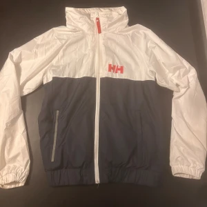 Helly Hansen vindjacka vit/marinblå - Snygg vindjacka från Helly Hansen i vit och marinblå med röd logga på bröstet och stort HELLY HANSEN-tryck på ryggen. 