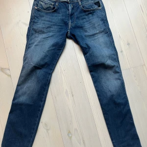 Blå Replay Anbass jeans - Snygga blå jeans från Replay, modell Anbass. Jeansen har klassisk femficksdesign, normal passform och är tillverkade i ett mjukt bomullsmaterial med lite stretch. Färgen är mörkblå med lätt slitning på låren och knäna för en nice look.