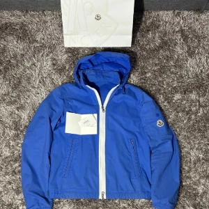 Blå vindjacka från Moncler med huva - Snygg blå vindjacka från Moncler med vit dragkedja och huva. Jackan är i storlek 1 och kommer med kvitto  