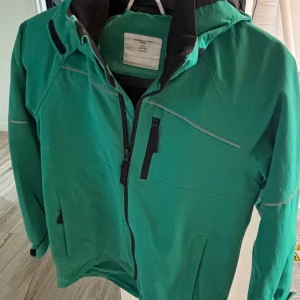Grön vindjacka från Polarn O. Pyret strl 134 - Snygg grön vindjacka från Polarn O. Pyret med svarta detaljer och dragkedjor. Jackan har huva, reflexdetaljer och praktiska fickor med dragkedja. Perfekt för blåsiga dagar och utomhusaktiviteter. Aningen sliten vid änden av dragkedjan, se bild. 