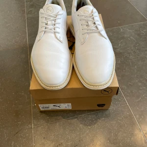 Puma Royale golfskor - Stilrena vita golfskor från Puma i skinn med klassisk snörning och rund tå. Skorna har greppvänlig sula med gummidobbar och diskreta detaljer i beige och brunt vid hälen. Perfekta för dig som vill ha en clean look på golfbanan. Skorna har används ett 10 tal gånger och ett extrem bra skick.            Det är bara att skriva om du har några frågor om skorna😃