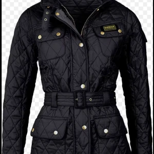Svart quiltad jacka från Barbour - Snygg svart quiltad jacka från Barbour International med bälte i midjan, guldiga knappar och flera fickor. Jackan har lång ärm, hög krage och klassisk Barbour-logga på bröstet. Perfekt för dig som gillar stilren och tidlös design.