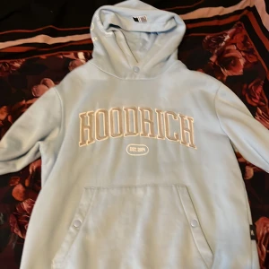 Ljusblå hoodie från Hoodrich S - Säljer en ljusblå hoodie från Hoodrich i storlek S. Tröjan har stor broderad logga framtill, tryck på ryggen och huva. Praktisk magficka med knappar och mjukt material som känns skönt mot huden. Perfekt för chill eller streetwear.