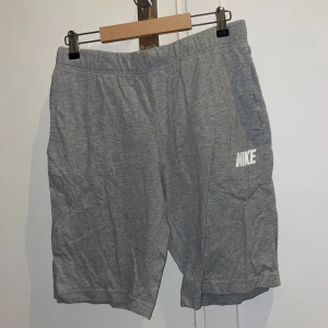 Grå shorts från Nike med logga - Gråa shorts från Nike med vit logga på vänster ben. De har elastisk midja och är tillverkade i mjuk bomull, vilket gör dem riktigt sköna. Klassisk, loose passform och fickor både fram och bak. Perfekta för chill eller träning.