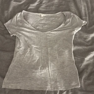 Ljusgrå t-shirt med v-ringning H&M - En enkel och snygg ljusgrå t-shirt från H&M i storlek M. T-shirten har v-ringad hals och korta ärmar, tillverkad i mjuk bomull som känns skön mot huden. Perfekt till jeans eller shorts och lätt att matcha med andra plagg.