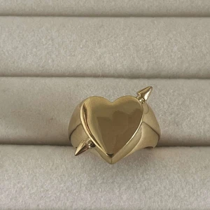 Guldfärgad ring med hjärta och spjut - Cool guldfärgad ring med ett stort hjärta i mitten och två spetsiga detaljer på sidorna. Ringen har en chunky design och är öppen baktill så den kan justeras. Perfekt för dig som gillar statement-smycken med edgy vibe.