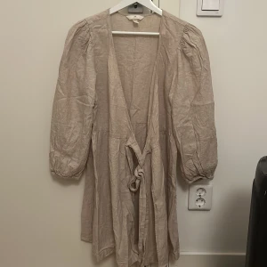Beige kort klänning från H&M i linneblandning - Säljer en beige kort klänning från H&M i linneblandning. Klänningen har puffiga långärmar, omlottmodell med knyt framtill och en avslappnad passform. Perfekt för dig som gillar en enkel och stilren look med naturliga material.