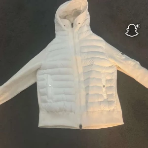 Vit Moncler dunjacka med stickade ärmar - Säljer en snygg vit Moncler dunjacka med stickade ärmar och huva. Jackan har quiltad front, dragkedja och två fickor med tryckknappar. Klassisk Moncler-logga på ärmen och coola detaljer. Perfekt för dig som vill ha en stilren och varm jacka.