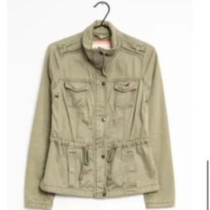 Olivgrön jacka från Hollister - Snygg och trendig olivgrön jacka från Hollister med dragsko i midjan och flera fickor framtill. Jackan har knappar och dragkedja samt en broderad logga på bröstet. Perfekt för lager-på-lager och chill stil. Jättesnygg till våren och hösten.