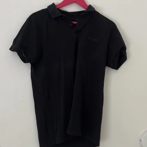 Svart kortärmad t-shirt från BOSS - Svart t-shirt från BOSS med klassisk krage och knappar framtill. Diskret BOSS-logga på bröstet. T-shirten är i mjuk bomull och har en stilren, enkel design som passar perfekt till jeans eller shorts.