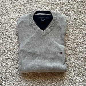 Grå tröja Tommy Hilfiger - Klassisk grå v-ringad tröja från Tommy Hilfiger med diskret logga broderad på bröstet. Tröjan är långärmad och har ribbade muddar vid ärmslut och nederkant. Tillverkad i mjuk bomull, perfekt för lager-på-lager under höst och vinter. Enda defekten är en linje rostfläckar från att ha hängt på en ställning. De är på baksidan av tröjan och syns på sista bilden. Men fläckarna syns knappt när man har på sig tröjan.