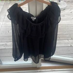Svart volangblus från Gina Tricot - Supersnygg svart blus från Gina Tricot med transparenta, vida ärmar och volangdetaljer längs ringningen. Blusen har en luftig och flowy känsla, perfekt för dig som gillar en trendig och romantisk stil.