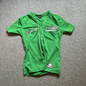 Grön fotbollströja World Wide MAN - Snygg grön fotbollströja från MAN med vit text 'World Wide' och broderad detalj på bröstet. Tröjan har korta ärmar, knappar framtill och vit piping längs kragen. Materialet är lätt och andas, perfekt för match eller träning. Fick den av mitt ex - så om du är en grabb eller tjej som vill ha denna - den är lite oversized är en M 