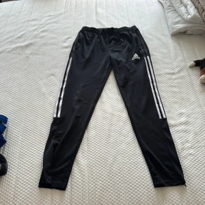 Svarta Adidas träningsbyxor! - Svarta träningsbyxor från Adidas med klassiska vita ränder längs sidorna och logga på låret. Byxorna har snörne så man kan knyta och dragkedja där nere för att kunna få på och av byxorna enklare.🥰