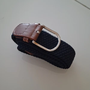 Mörkblått flätat bälte med skinn - Snyggt mörkblått flätat bälte med detaljer i brunt skinn och silvrigt spänne. Bältet har en klassisk design med vävd struktur och passar perfekt till jeans eller chinos. Skinnet har vita sömmar som ger en schysst kontrast mot det mörka bandet.