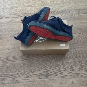 Mörkblå sneakers från Louboutin, strl 45 - Säljer ett par mörkblå sneakers från Louboutin i mocka med klassisk röd sula. Skorna har snörning och rund tå, samt en stilren design med diskreta detaljer. Perfekta för dig som vill ha något exklusivt och snyggt på fötterna. Dem är använda nästan halva sommaren, man kan lätt tvätta dem så de ser nya ut om man orkar det.