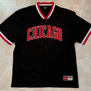 Svart Chicago Tröja med Knappar - Snygg svart tröja med knappar från FSBN i storlek S med CHICAGO tryckt i rött och vitt över bröstet. Tröjan har korta ärmar och knappar framtill samt röda och vita detaljer på krage och ärmslut. Frakt tillkommer!