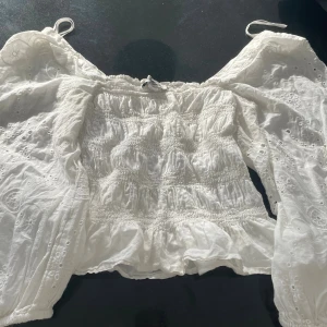 Vit blus med puffärm från Hampton Rep - Supersöt vit blus från Hampton Rep i storlek XS. Blusen har smockad kropp, vida puffärmar och broderade detaljer med hålmönster. Offshoulder-modell med resår upptill och nertill på ärmarna. Perfekt för en somrig look!