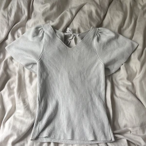 Ljusgrå ribbad topp med puffärm - Superfin ljusgrå ribbad topp med korta puffärmar och v-ringning. Toppen har knytning i nacken och är tillverkad i ett mjukt, stretchigt material. Perfekt för dig som gillar en enkel men ändå söt stil.