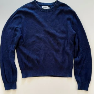 Mörkblå stickad tröja från East West - En stilren mörkblå stickad tröja från East West med rund halsringning och långa ärmar. Tröjan har en ribbad struktur och en klassisk passform som funkar till många olika looks.