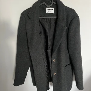 Mörkgrå kappa från Noisy May - Snygg mörkgrå kappa från Noisy May i ullmix med klassisk krage och två knappar framtill. Fodrad insida och tre dekorativa knappar vid ärmslutet. Perfekt till hösten och för dig som gillar tidlösa plagg. Använd fåtal gånger, inga defekter.