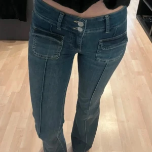 Blå bootcut jeans med fickdetaljer - Snygga blå jeans med bootcut passform och markerade sömmar längs benen. Byxorna har dubbla knappar i midjan, stora fickor framtill med dragkedjedetaljer och klassiska bakfickor. Perfekta för dig som gillar en cool och trendig look.