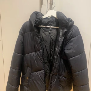 Svart pufferjacka från Vero Moda - Säljer en svart pufferjacka från Vero Moda med luva och dragkedja. Jackan är quiltad och har en höstligt  vibe, perfekt för kalla dagar. Fickor med knappstängning på sidorna och hög krage för extra värme.