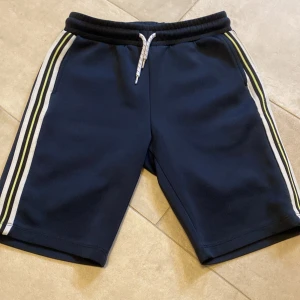 Mörkblå shorts med ränder från Lindex - Hej! Jag säljer ett par mörkblå shorts från Lindex med vita och ljusgröna ränder längs sidorna. De har elastisk midja med snörning och två fickor framtill. Den är lite  lite nopprig på vänstra sidan men det syns knappt och påverkar ingenting. Perfekta för en sportig och avslappnad stil. Tveka inte att höra av dig vid frågor!
