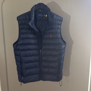 Mörkblå dunväst från Polo Ralph Lauren - Snygg mörkblå dunväst från Polo Ralph Lauren med klassisk quiltad design och dragkedja framtill. Västen har två fickor med dragkedja och den ikoniska röda loggan broderad på bröstet. Perfekt för lager-på-lager-stil under kyliga dagar. Storlek M in köptes ny för 2500kr i princip aldrig använd 10/10