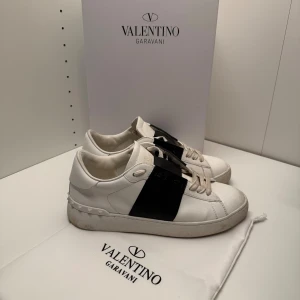 Valentino opens  - Snygga sneakers från Valentino Garavani i vitt skinn med bred svart läderdetalj över sidan. Klassisk rund tå, platt sula och coola nitar på hälen. Skorna har snörning och kommer med originalpåse och box. Perfekt för dig som gillar exklusiv streetstyle. Allt og nedkommer som box, skopåse, påse och kuvert. Pm vid funderingar.