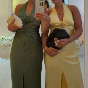 Olivgrön långklänning med halterneck - Säljer en olivgrön långklänning med halterneck och snygg rosettdetalj framtill. Klänningen har en slits framtill och är figurnära med en elegant, glansig finish. Perfekt för dig som vill sticka ut med en stilren och trendig look.