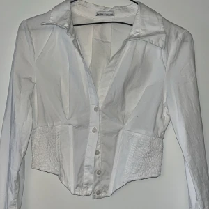 Vit croppad skjorta från Berthia - Snygg vit skjorta från Berthia med croppad passform och smockade partier i midjan. Skjortan har klassisk krage, långa ärmar och knäppning framtill. Perfekt för dig som vill ha en stilren men ändå unik look.