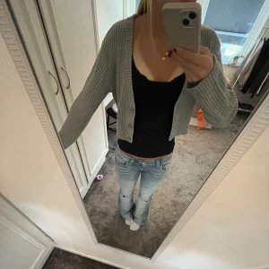 Grå ribbad stickad kofta - Snygg grå stickad väst med ribbad struktur och v-ringad hals. Modellen är croppad och har knappar framtill. Perfekt att slänga över en topp för en chill och trendig look.