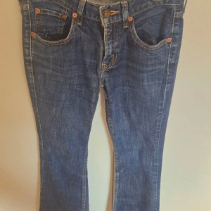 Levi's 501 blå jeans bootcut - Klassiska blå Levi's 501 jeans med bootcut-snitt. Jeansen har fem fickor, orange kontrastsömmar och den ikoniska röda Levi's-taggen på bakfickan. Tillverkade i slitstarkt bomullsjeansmaterial och har en rak passform upptill som går ut något vid benslutet.