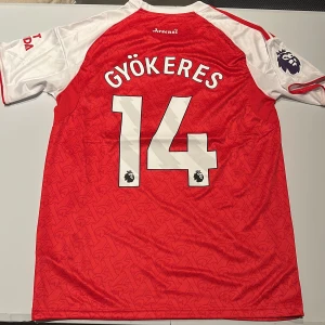 Arsenal Gyökeres 14 Adidas matchtröja - Säljer en officiell Arsenal fotbollströja från Adidas med Gyökeres och nummer 14 på ryggen. Tröjan är röd med vita ärmar, har Premier League-märke på ärmen och diskret mönster i tyget. Materialet är lätt och andas, perfekt för match eller träning. Replika, endast handtvätt gäller!