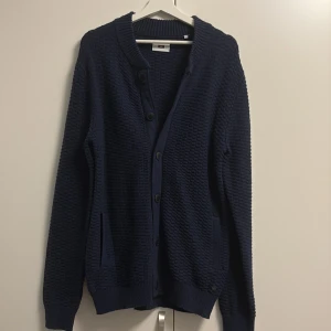 Mörkblå stickad kofta från WE - Mörkblå stickad kofta från WE med grov struktur, ribbade muddar och knäppning framtill. Långärmad modell med två fickor och klassisk v-ringning. Perfekt för lager-på-lager och chill vibes. Stor i storleken 